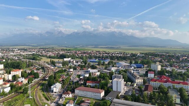 Drone flying backward and ascending over Poprad, showing the cityscape and the majestic High Tatras on a bright clear day.