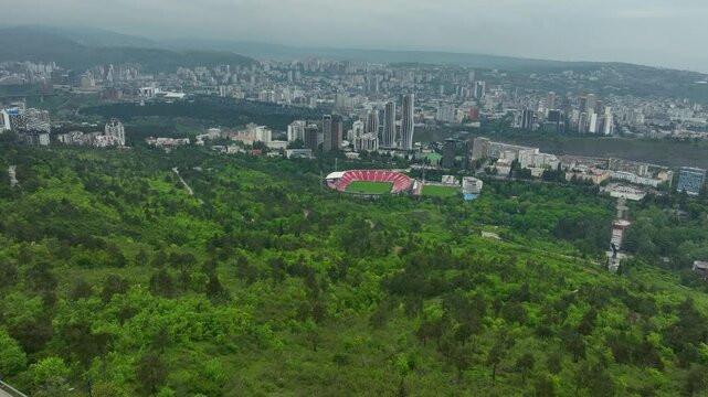 Aerial view of Tbilisi city framed by green hills and the red roof of the Dinamo Arena stadium visible amid the urban landscape.