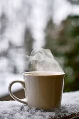 Steaming ceramic mug standing on snowy surface outdoors with blurred winter forest background, hot beverage emitting visible vapor, closeup capturing warmth and contrast