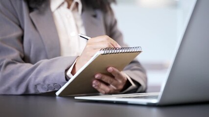 Close up of female hand taking notes in notebook while using laptop at work desk at workplace in office. African American businesswoman writes with a pen during a remote business meeting or conference - Powered by Adobe