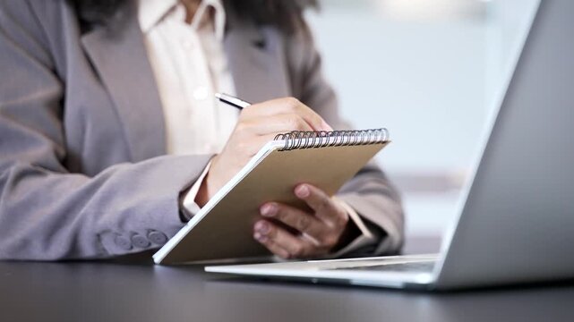 Close up of female hand taking notes in notebook while using laptop at work desk at workplace in office. African American businesswoman writes with a pen during a remote business meeting or conference