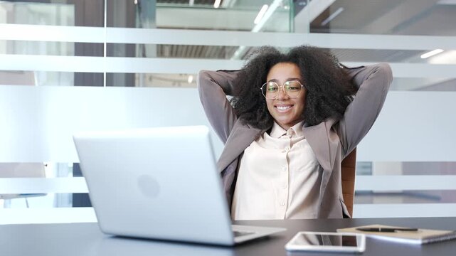Happy african american businesswoman finished work on laptop while sitting at workplace in office. Smiling satisfied female manager puts her hands behind her head, leaning back in chair. Work done