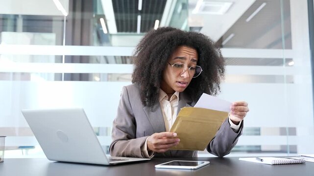 Disappointed african american businesswoman reading letter with bad news while sitting in business office. Upset female manager is depressed by unpleasant notification, shocked after opening envelope