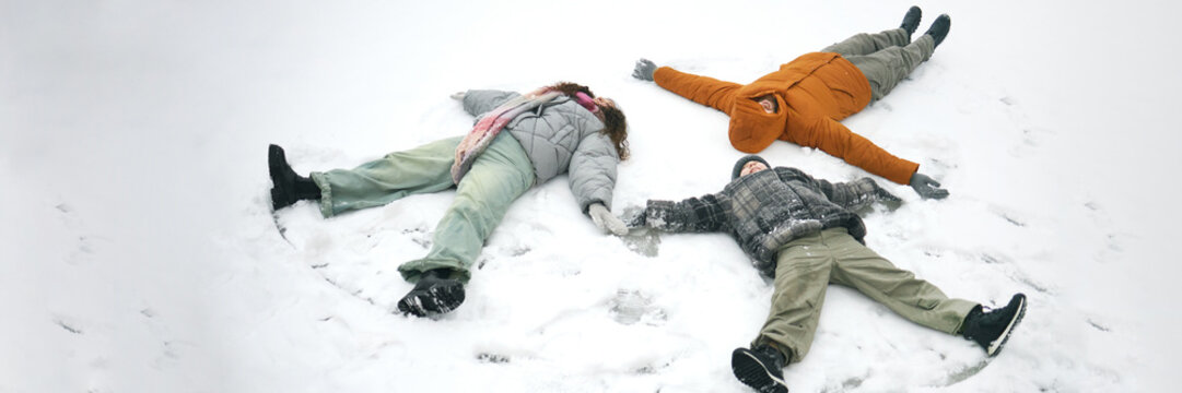 Header of family of parents and kid lying on snow creating snow angels, arms and legs spread wide, winter clothing visible, overhead view capturing playful outdoor winter activity together