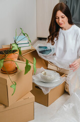 Portrait of woman wearing white t-shirt sitting on the floor surrounded with carton boxes and packing plates and dishes. Relocating, buying new house. Pack personal stuff from carton boxes. 