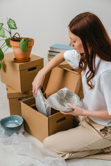 Portrait of woman wearing white t-shirt sitting on the floor surrounded with carton boxes and packing plates and dishes. Relocating, buying new house. Pack personal stuff from carton boxes.