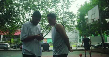 Latin Hispanic man of African descent and Hispanic friend looking at smartphone on basketball court, teamwork and camaraderie during urban sports practice outdoors - Powered by Adobe