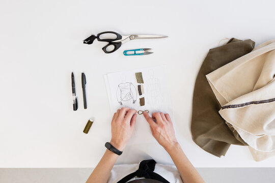 Directly above view of unrecognizable female tailor choosing right fabric for garment while standing by table with sketch