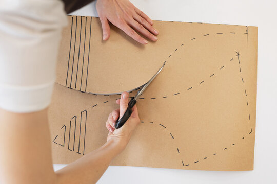 Top view of unrecognizable female tailor standing by table, holding scissors and cutting out patterns for new clothing