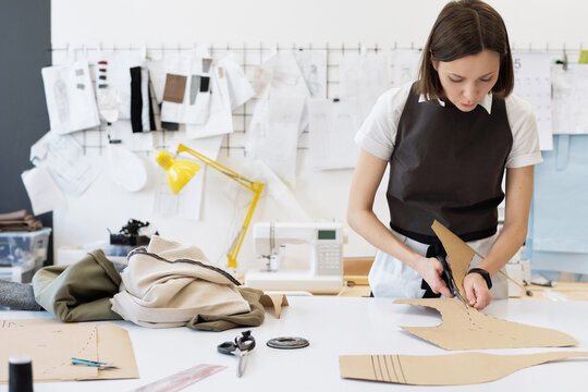 Female fashion designer cutting out templates for garment while standing by table in tailor workshop