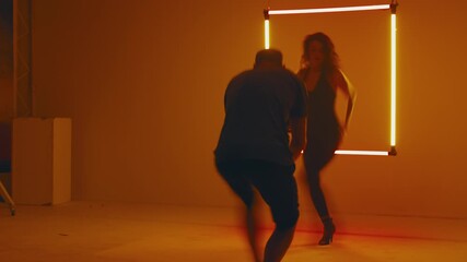 In studio setting two women dance in front of glowing neon square, their movements enhanced by the rotating fan’s motion with the vibrant light outlining their silhouettes - Powered by Adobe