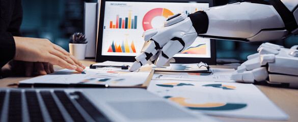 A robotic hand collaborates with a human in analyzing business data using charts and graphs in a...