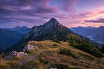 Fototapeta premium Serene mountain landscape with a winding path through grass and rocks, framed by shrubs, showcasing majestic forested peaks under a warm sunset sky, evoking tranquility and nature's beauty