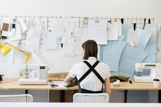 Rear view of unrecognizable female tailor sewing garment in atelier while sitting at table on background of multiple templates