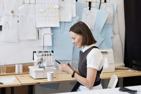 Side view of young Caucasian female tailor sitting by table with sewing machine, using smartphone and smiling