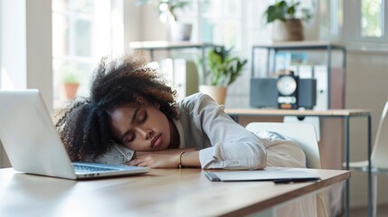 Obraz premium Young African woman with curly hair resting on a desk in a modern office. A laptop and notebook are visible on the table. Indoor setting with plants in the background.