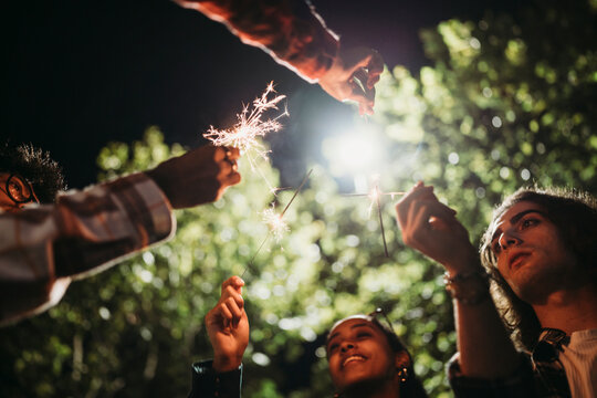 from below crop of people doing a party in casual clothes with burning sparklers outdoors at night
