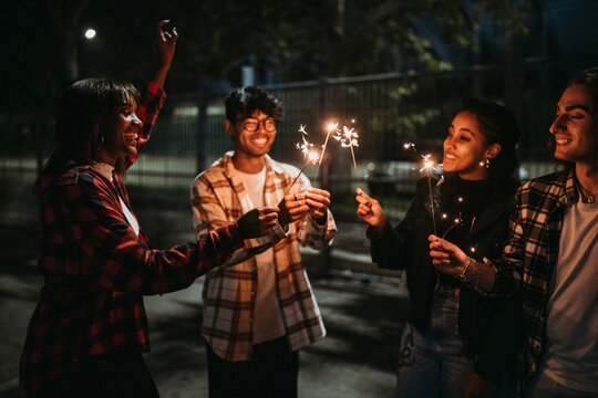 Positive young multiracial friends doing a party in casual clothes smiling and looking at burning sparklers while standing on city street at night