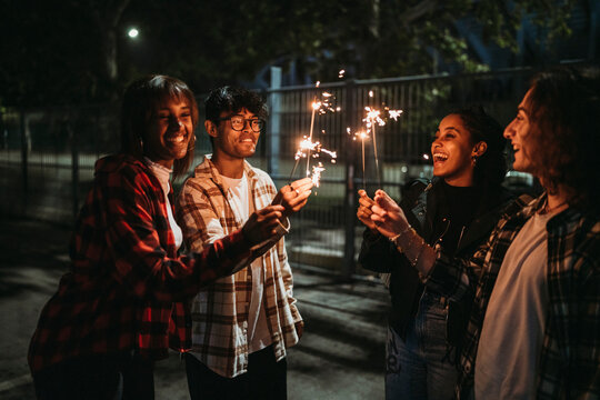 Positive young multiracial friends doing a party in casual clothes smiling and looking at burning sparklers while standing on city street at night