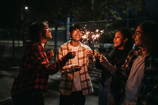 Positive young multiracial friends in casual clothes smiling and looking at burning sparklers while standing on city street at night
