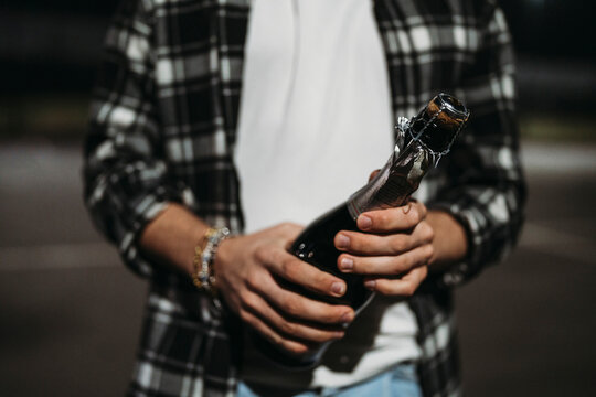 Crop hand of man in checkered shirt holding bottle of champagne while standing on blurred background of street at night