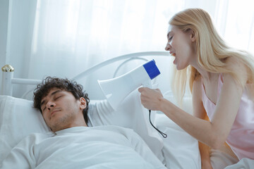 A white woman screams into a megaphone at her sleeping husband, trying to wake him up in the...