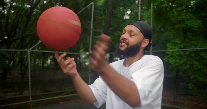 African descent man wearing durag spinning basketball on finger at outdoor court, expressing skill, confidence, and relaxed joy in casual urban sports moment