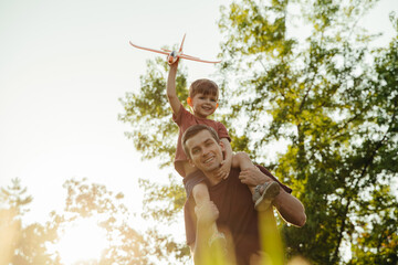 Father and young son enjoy bonding time in nature, with the child excitedly holding a toy airplane while sitting on dad's shoulders. Portrait of happy father with playful son outdoors. Space for text