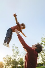 Joyful father playing with his smiling young son, tossing up and catching him in a park at sunset....