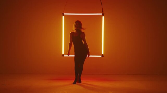 Woman in tight black sexy outfit and high heels dances modern dance against the backdrop of a neon square light.