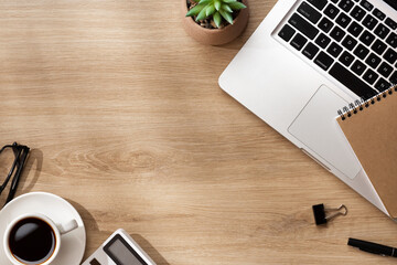 A morning workspace with a notebook, laptop, coffee, calculator, plant, pen and glasses on a wooden surface, softly lit sunlight from the window.