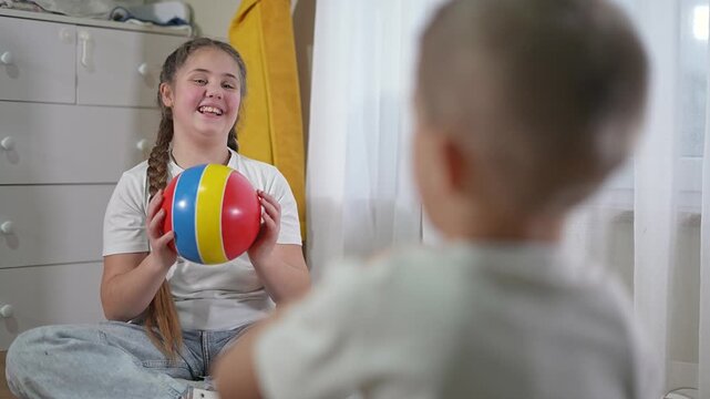 Boy smiles holding toy ball and looking at friend. Child stays seated as game continues indoors. Playful exchange between child and friend adds joy to quiet activity. Boy waits for next throw.