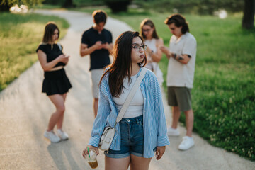 Obraz premium A young woman stands thoughtfully in a park while her friends use their phones, highlighting the theme of digital interaction and its effect on social dynamics in natural spaces.