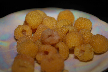 This detailed macro shot captures a pile of ripe yellow raspberries placed on a light iridescent plate, against a dark background.