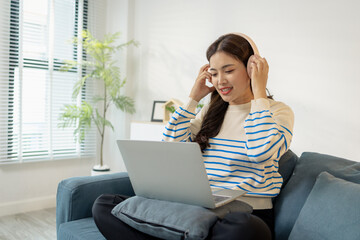 Fototapeta premium A young woman wearing headphones sits comfortably on the sofa, smiling while working on her laptop. She looks focused and happy, representing modern lifestyle and remote work.