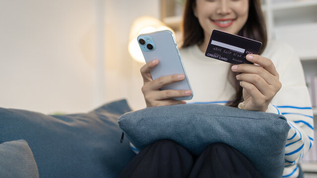 A young woman is sitting on a sofa holding a smartphone and credit card, smiling as she makes an online transaction. She looks relaxed and confident while shopping at home.