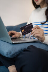 A young woman sits comfortably on a sofa using a laptop while holding a credit card, appearing to make an online payment. She smiles with satisfaction, reflecting ease and convenience.