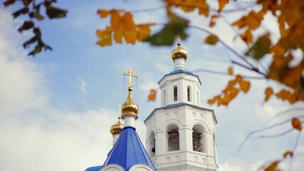 Golden leaves frame blue domed orthodoxy church towers against cloudy sky, yellow autumn leaves in foreground.