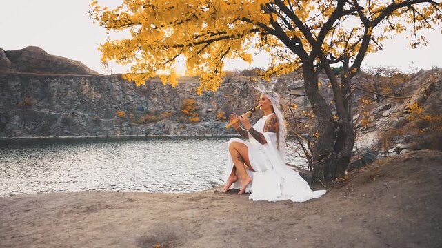 fantasy woman fairy faun plays a flute, sitting under tree creating magical melody fairytale music. Myth girl with deer antlers on head, blonde hair long white dress fly in wind. river rock sun light