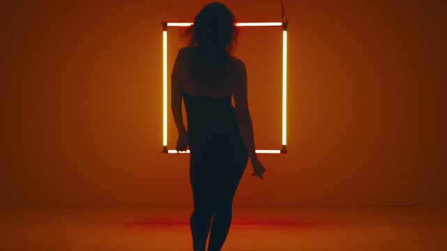 Woman silhouette steps away from the camera, taking her place before the glowing neon square as she starts to dance