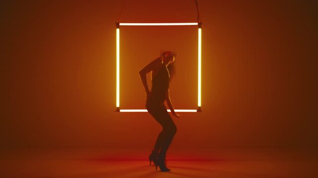 Woman in tight black sexy outfit and high heels dances modern dance against the backdrop of a neon square light