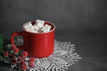 Red mug with hot chocolate and marshmallows on a dark background. A winter composition with berries and a knitted napkin, a symbol of comfort and celebration.