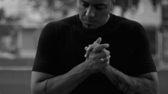 Hispanic man of African descent standing outdoors looking down with worried expression and clasped hands, emotional black and white portrait conveying anxiety and reflection