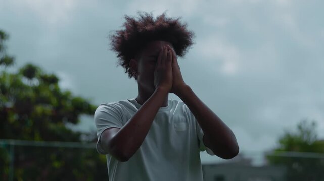Latin Hispanic man of African descent standing outdoors with hands pressed in prayer, seeking inner peace and strength under cloudy sky, expressing faith and emotional surrender