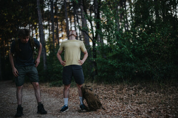 Fototapeta premium Two men pause during a forest hike. One gazes upward while the other bends, with a backpack on the ground, capturing outdoor exploration and friendship amid autumn leaves.