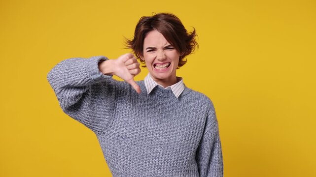 Abhorred disgusted sad young Caucasian woman she wear grey knitted sweater white shirt casual clothes pointing thumb down dislike gesture isolated on plain yellow color background. Lifestyle concept