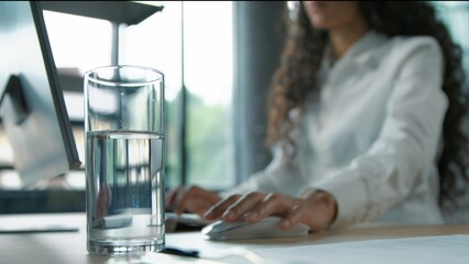 Close up glass of still water standing on table in business office blurred view unrecognizable woman businesswoman female girl worker working with computer internet typing chatting online aqua balance