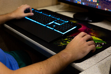 Close up of hand on gaming mouse, with backlit keyboard glowing in blue, showcasing colorful gaming mousepad. The screen displays game, highlighting the immersive experience