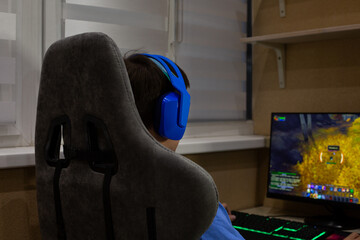 Young gamer wearing blue headphones sits at desk, playing an online video game on computer. The image captures the immersive experience of modern gaming, with vivid on screen action