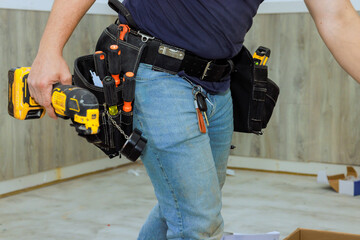 Construction worker is actively using power drill in home under renovation, using tool belt...